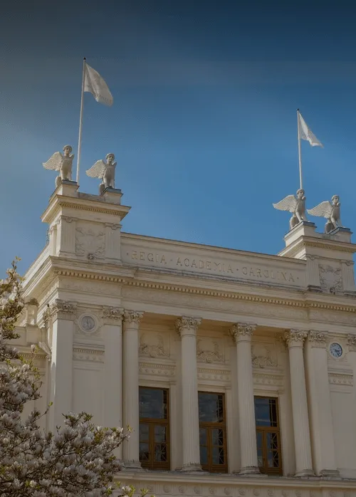 Classical building façade with four columns, winged statues on top, and flags against a clear blue sky.