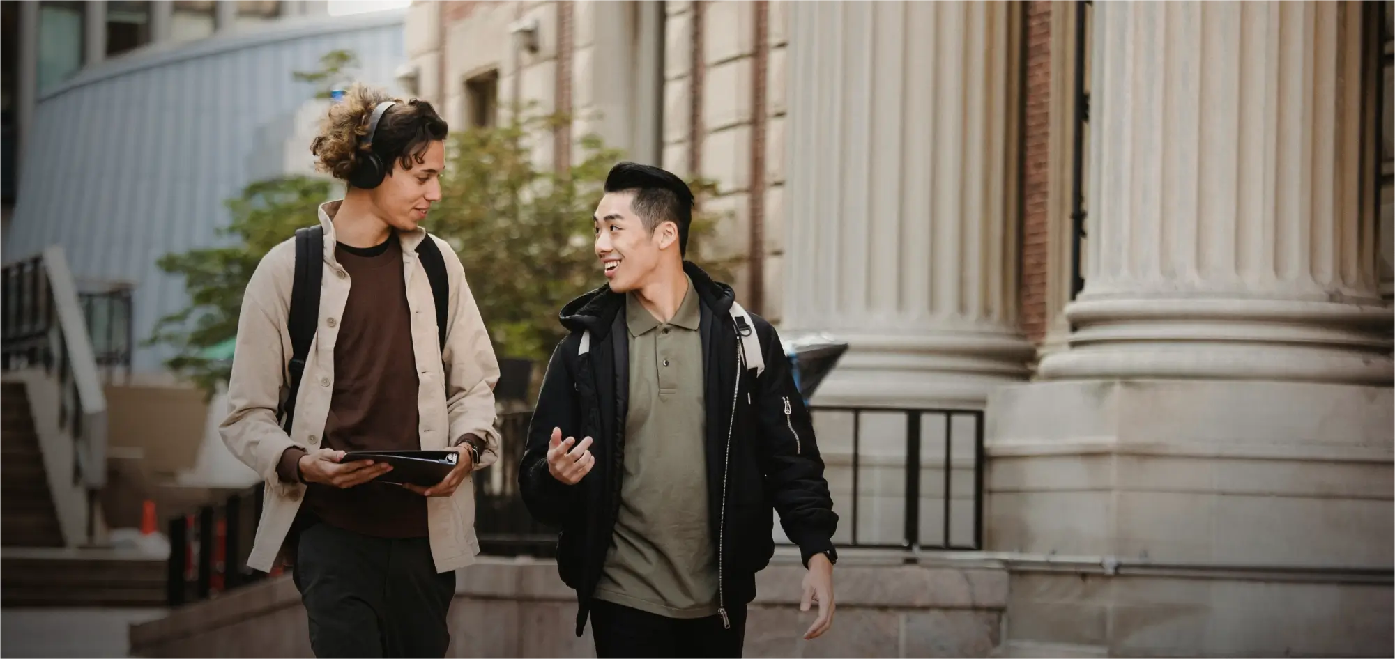 A photograph of two young male students walking and talking on a college campus sidewalk.