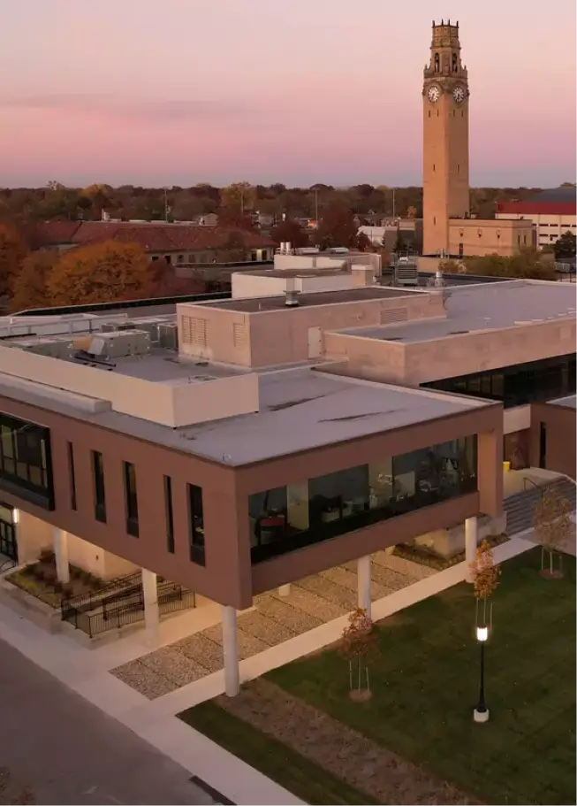 An elevated view of a university campus at sunset or sunrise, characterized by a soft pink and purple sky. In the foreground is a modern, two-story building with a flat roof, large windows, and a brown and white facade. In the background, rising above the surrounding buildings and trees, is a tall, historic-looking brick clock tower or bell tower. The ground level shows paved walkways and green lawn.