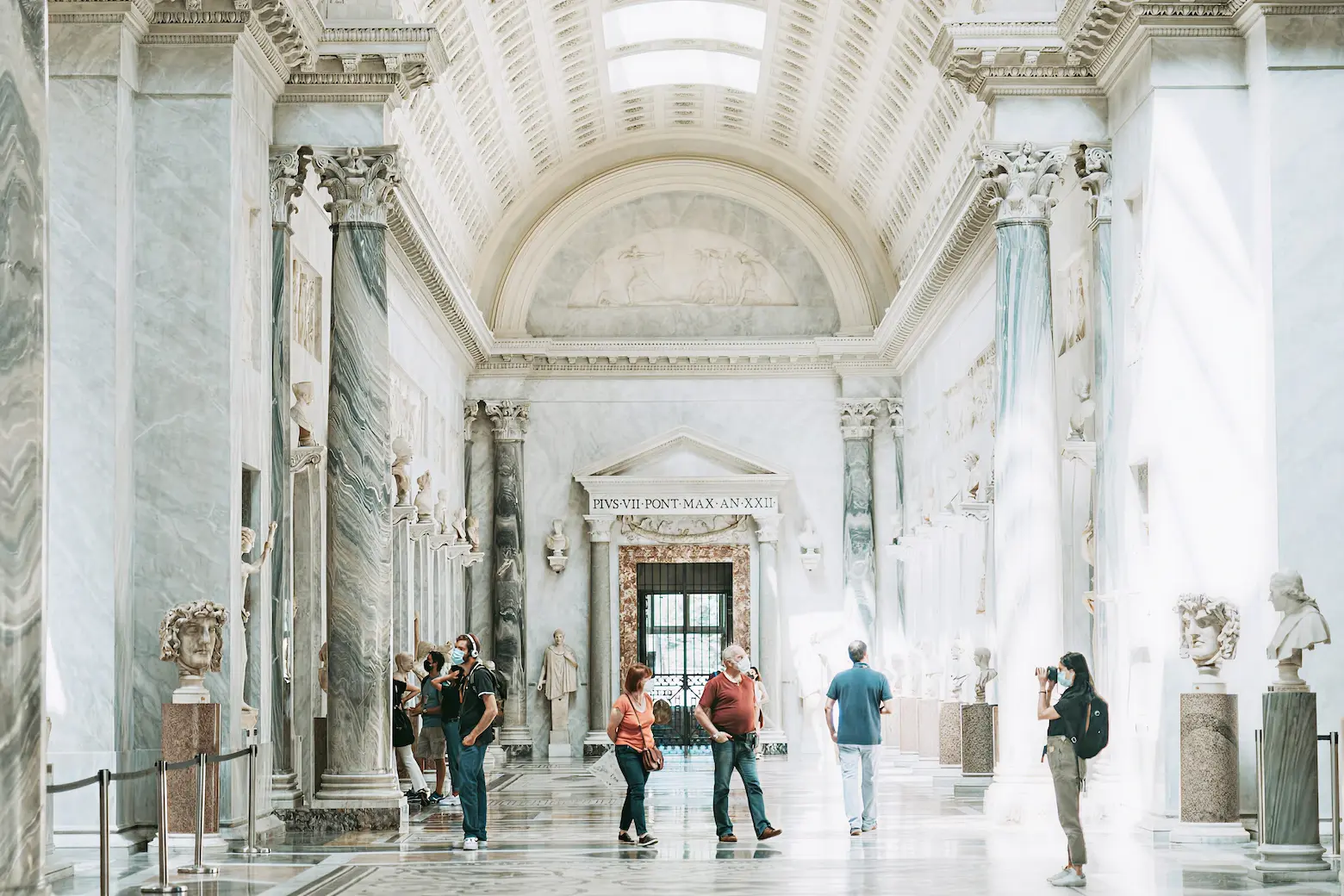 Visitors wearing masks observe classical sculptures in a marble gallery with tall columns and vaulted ceilings.