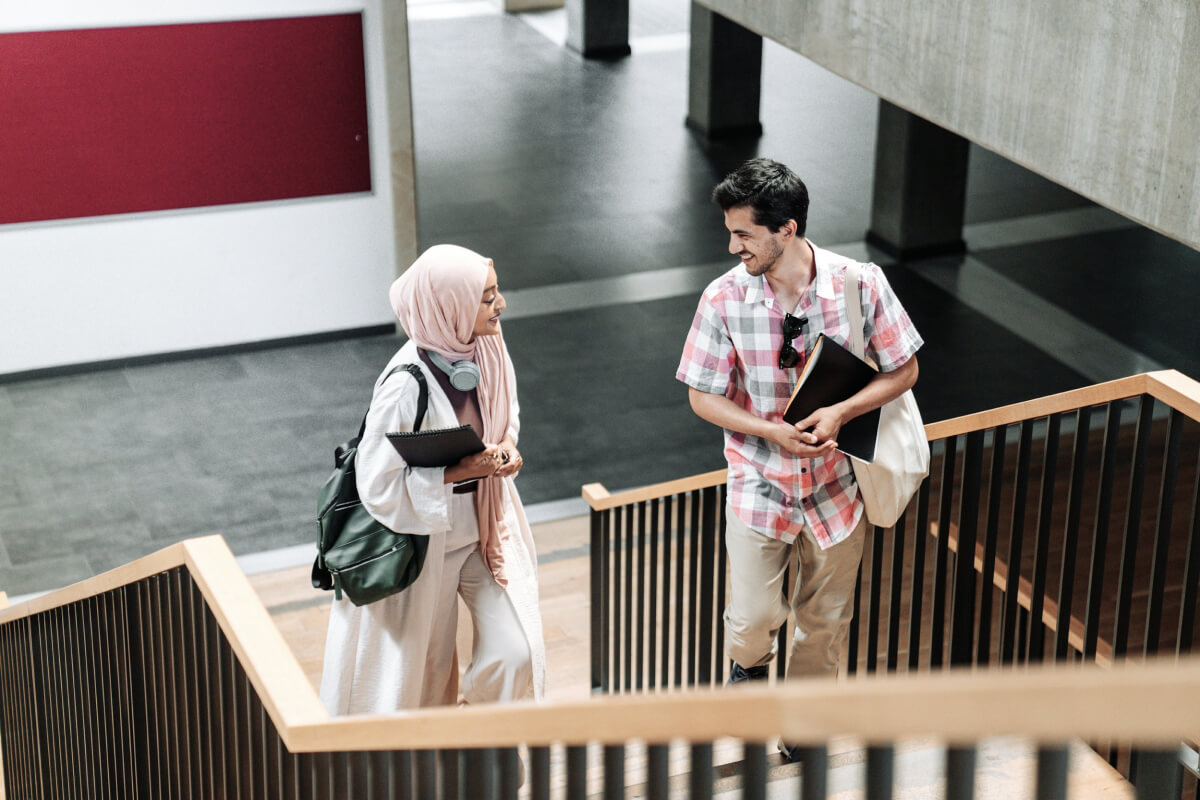 Two students, a woman in a light pink hijab and a man in a checkered shirt, walking up stairs and smiling at each other indoors.