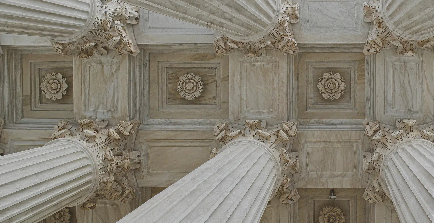 Dramatic upward view of a coffered, light-colored marble ceiling featuring carved rosettes in the recessed panels. The ceiling is framed and supported by large, fluted classical columns with highly ornate Corinthian or Composite capitals carved with intricate acanthus leaves.